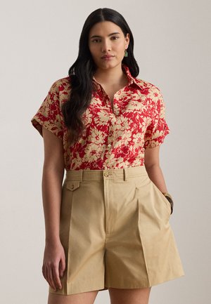 Femeie cu păr lung și închis la culoare, purtând o cămașă roșie cu model floral, cu mâneci scurte și nasturi, băgată în pantaloni scurți bej cu talie înaltă, având mâinile în buzunare.