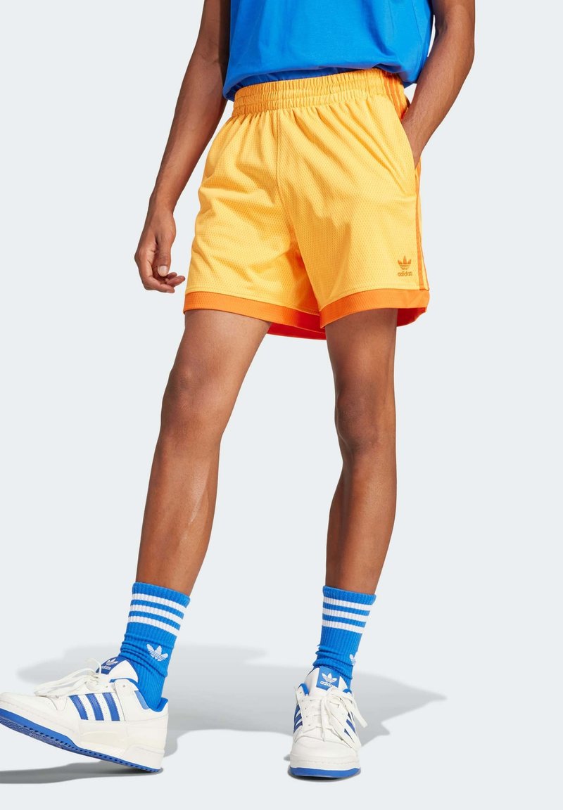 Orange mesh shorts with an elastic waistband, featuring contrast stitching and a side logo. Paired with blue striped socks and white sneakers.