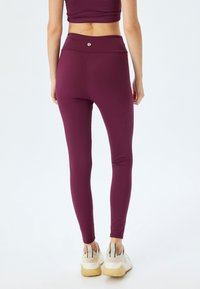 Bordeauxrood leggings met een hoge taille, een gladde textuur en minimale branding. Het model draagt witte en beige sneakers.