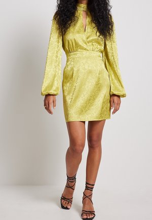 Robe mini verte citron à motif floral avec de longues manches volumineuses, encolure en trou de serrure et taille élastique. Associée à des sandales noires à lanières.