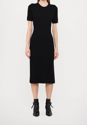 Femme portant une robe midi noire cintrée à manches courtes, associée à des bottines noires avec fermeture éclair et détails de boucles.