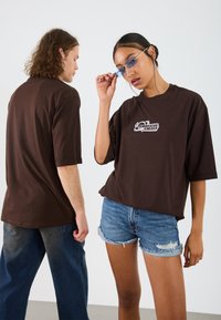 Camisa de algodón marrón de gran tamaño con mangas cortas, que presenta un logo gráfico. Combínala con pantalones cortos de mezclilla azul. Textura suave, corte casual.