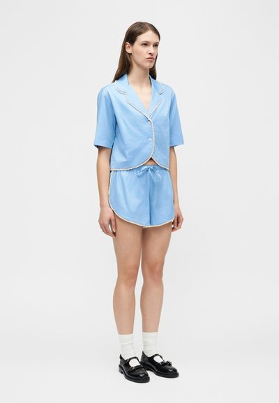 Femme portant une chemise à manches courtes bleu clair boutonnée avec un liseré blanc et un short assorti, accompagnée de chaussettes blanches et de mocassins noirs.