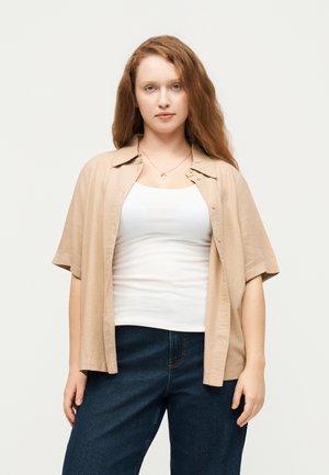 Tânără cu păr lung roșcat, purtând o cămașă bej deschisă peste un maieu alb și blugi albastru închis, stând în fața unui fundal simplu.