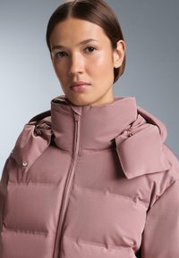 Puhana jakna u nježnoj ružičastoj boji, s visokim ovratnikom, jedinstvenim detaljem s gumbima te glatkom teksturom, sa zatvaračem i suptilnim sjajem.