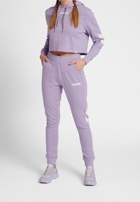 Lila verkürzter Hoodie mit weißem Logo; passende lila Jogginghosen mit weißen Akzenten und Taschen, kombiniert mit helllila Sneakers.