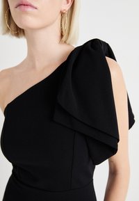 Musta mekko, jossa on yhden olkapään malli, sisältäen suuren, taitetun kankaan yksityiskohdan. Silkkinen tekstuuri ja vartaloa myötäilevä leikkaus. Kultaiset renkaat. 