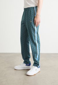Pantalons de survêtement verts à motifs avec des rayures latérales bleues et blanches, taille élastique et accent avec le logo Lacoste. Portés avec des baskets blanches.
