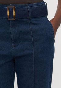 Mørkeblå denimbukser med en glat overflade, der har et bredt bælte med en rektangulær brun spænde og synlige sømdetaljer.