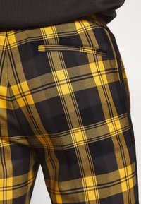 Pantalones de cuadros amarillos y negros con tela suave, que cuentan con un bolsillo trasero y un ajuste entallado. Patrón de cuadrícula audaz con líneas contrastantes.