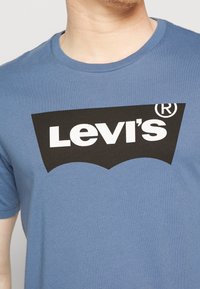 Homme portant un t-shirt bleu avec un logo Levi's noir et un texte blanc centré sur la poitrine.