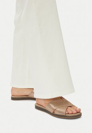 Bronzefarbene gewebte Slide-Sandalen mit Kreuzriemen, flacher Sohle und Gummiaußensohle, kombiniert mit weißen ausgestellten Hosen, die eine entspannte Passform zeigen.