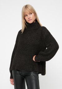 Brauner Rollkragenpullover mit strukturiertem Strick, übergroßen Ärmeln und einer lässigen Passform, kombiniert mit schwarzen Kunstlederhosen.