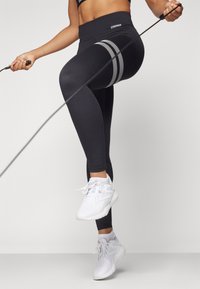 Black high-waisted leggings with striped detail; paired with white athletic shoes. The leggings feature a smooth texture and fitted design.