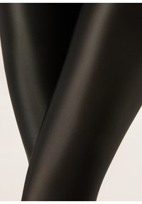 Primo piano di gambe incrociate che indossano leggings neri lucidi e lisci su uno sfondo chiaro e uniforme.