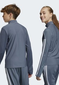 Giubbotti sportivi grigi con un design testurizzato, colletto alto e strisce bianche lungo le maniche, indossati da due modelli visti da dietro.