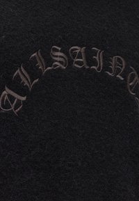 Black textured fabric with the word "BALMAIN" embroidered in dark brown Gothic-style letters in a curved arrangement.