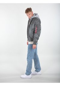 Giubbotto grigio puffo con cappuccio, chiusura a zip, polsini a coste e dettaglio di etichetta rossa. Indossato sopra una maglietta grigia chiara con jeans azzurri chiari.