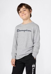 Uśmiechnięty chłopiec w szarej koszulce Champion z długim rękawem i czarnych dresach Champion, z jedną ręką w kieszeni, stojący na tle białym.