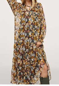 Nainen, joka pukeutuu pitkään, läpikuultavaan kukkapukuun, jossa on keltaisia, valkoisia ja marjapuuron värisiä kukkia, nostaa hameen paljastaakseen vihreät polviin ulottuvat saappaat.