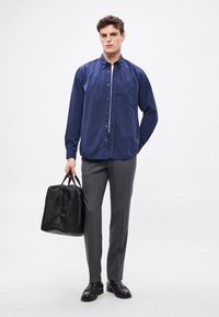 Cămășe cu nasturi bleumarin cu buzunar, pantaloni gri croiți și pantofi din piele neagră. Ține o geantă duffel din nylon negru.