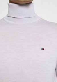 Jasnofioletowy sweter z golfem wykonany z prążkowanej dzianiny, z małym, haftowanym logo w czerwonym, białym i niebieskim kolorze na klatce piersiowej.