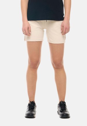 Femme portant un short beige, un haut noir et des chaussures de sport noires, debout avec les mains détendues le long du corps sur un fond blanc.