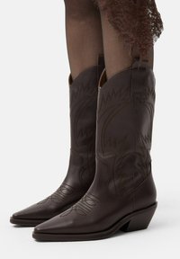Brūnas ādas cowboy zābaki ar norādītām pirkstu daļām, sarež�ģītu šuvju un zemu papēdi. Iezīmējas ar izliekta augšējo malu un vilkšanas cilpām sānos.