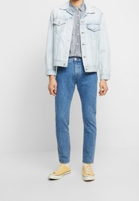 Ljusblå denimjacka med två bröstfickor, randig skjorta med knappar, blå jeans och gula sneakers med vita sulor och snörningar.