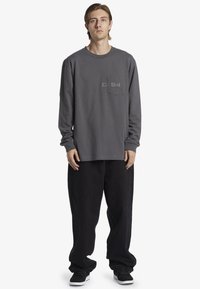 Grå långärmad skjorta med en framficka som har ett subtilt tryck. Kombinerad med löst sittande svarta byxor och sneakers.