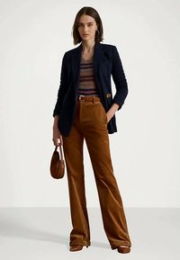 Ženska v temno modrem blejzerju, s črtasto puloverjem in rjavimi širokimi hlačami, drži majhno rjavo torbico in stoji pred enobarvno ozadjem.