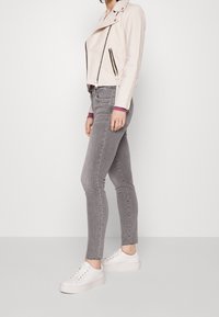 Vrouw in een lichtroze cropped jack, grijze skinny jeans en witte platform sneakers, staand tegen een effen witte achtergrond.