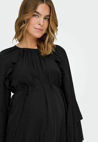 El vestido de maternidad negro presenta un diseño fluido con acentos de volantes y una cintura atada, hecho de una tela texturizada. Mangas largas con volantes en capas.
