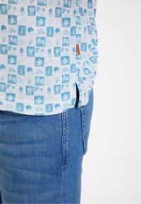 Srajca s kratkimi rokavi v svetlo modri barvi z geometrijskim vzorcem različnih oblik. Tkanina je teksturirana. Ujemanje z modrimi kavbojkami, ki prikazujejo ozek kroj.