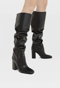 Jambes portant des bottes en cuir noires, slouchy, montantes jusqu'au genou avec des talons carrés, se tenant contre un fond clair et uni.