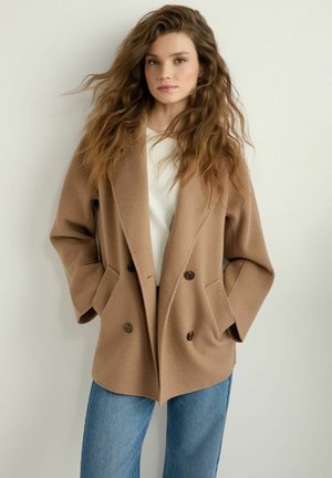 Młoda kobieta z długimi falującymi włosami, ubrana w beżowy płaszcz z podwójnym zapięciem, biały top i niebieskie dżinsy, stojąca na jednolitym tle.