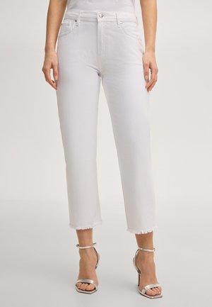 Kobieta w białych krótkich jeansach z postrzępionymi końcami i srebrnych sandałach na wysokim obcasie, stojąca na jednolitym tle.