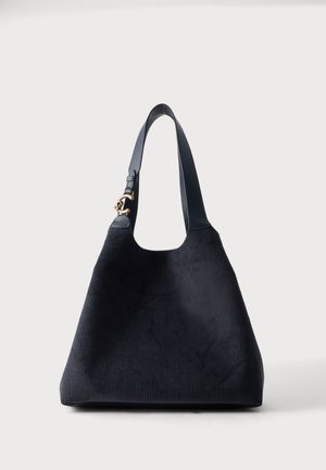 Borsa tote in velluto a coste nero con una larga tracolla singola e dettaglio gancio tono oro su sfondo bianco.
