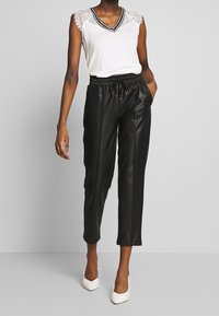 Biała bezrękawnikowa bluzka z koronkowym wykończeniem i dekoltem w kształcie V, zestawiona z czarnymi, krótki spodniami z sztucznej skóry i białymi, spiczastymi obcasami.