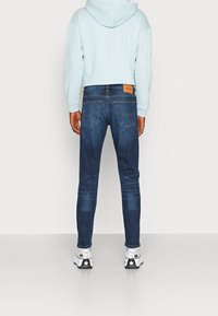 Slim-fit mörkblå jeans med en subtil blekt textur, utrustade med en läderlapp på midjan och kontrasterande sömmar. Bärs med en ljusblå hoodie.