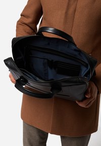 Schwarze Ledertasche mit doppelten Griffen, Reißverschluss und einem gefütterten Innenraum mit mehreren Taschen und Fächern.
