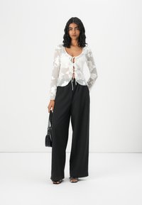 Transparent white long-sleeve top with floral embroidery, tie front detail, paired with high-waisted black wide-leg pants and black handbag.