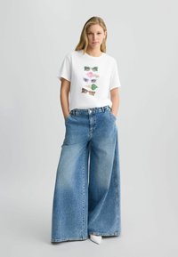 T-shirt blanc avec un graphique de lunettes de soleil colorées, associé à un jean en denim bleu à jambe large. Le mannequin se tient avec les mains dans les poches contre un fond uni.