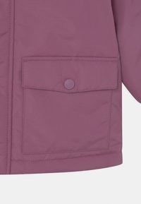 Détail d'une veste en duvet violette avec une poche frontale carrée dotée d'une fermeture à bouton-pression et d'un tissu lisse et texturé.