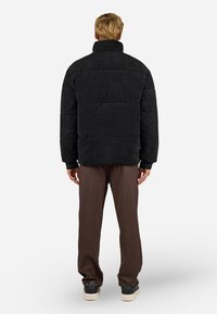 Sort corduroy pufferjakke med høj krave, quiltet design og blødt materiale. Modellen har brune bukser og sneakers på.