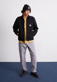 Chaqueta negra con cremallera y parche de logo, camiseta amarilla debajo, pantalones grises y zapatos negros de cordones con suela gruesa. Gorro de punto negro.