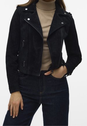 Kvinde iført en sort ruskind bikerjakke med sølv lynlåse over en beige rullekrave og mørkeblå jeans, hånd i lommen.