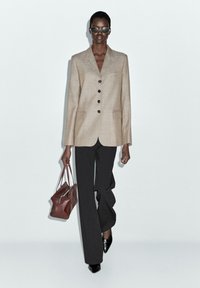 Beige tailored blazer with black buttons, paired with black wide-leg trousers. Brown leather bag and round sunglasses complete the look.