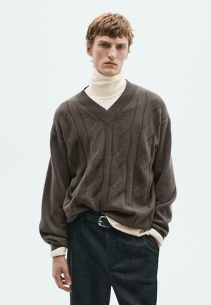 Brown, textured knit sweater with a V-neck and cable patterns, layered over a cream turtleneck. Worn with dark trousers and a black belt.