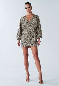 Zebra raksta mini kleita bēšā un melnā krāsā ar ietinamu dizainu, plūstošām piedurknēm un pieguļošu vidukļa daļu. Kopā ar zelta krāsas augstpapēžu sandales ar siksniņām.